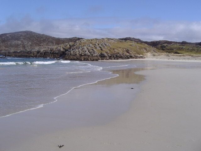 A sandy beach with gentle waves, rocky hills in the background, and a clear sky.