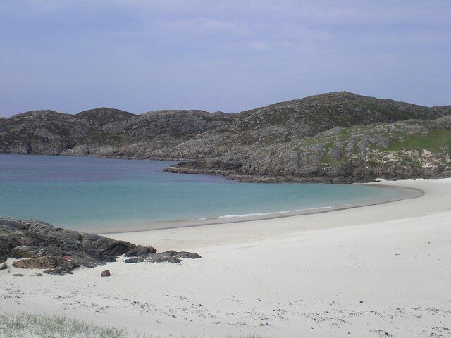 A beach with white sand, clear turquoise water, and rocky hills in the background.