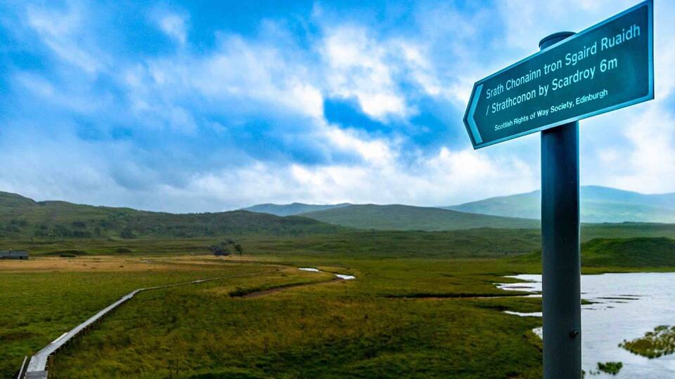 A signpost with a view of a wetland landscape, a wooden bridge, and distant hills.