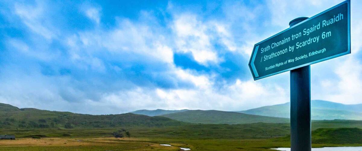 A signpost with a view of a wetland landscape, a wooden bridge, and distant hills.