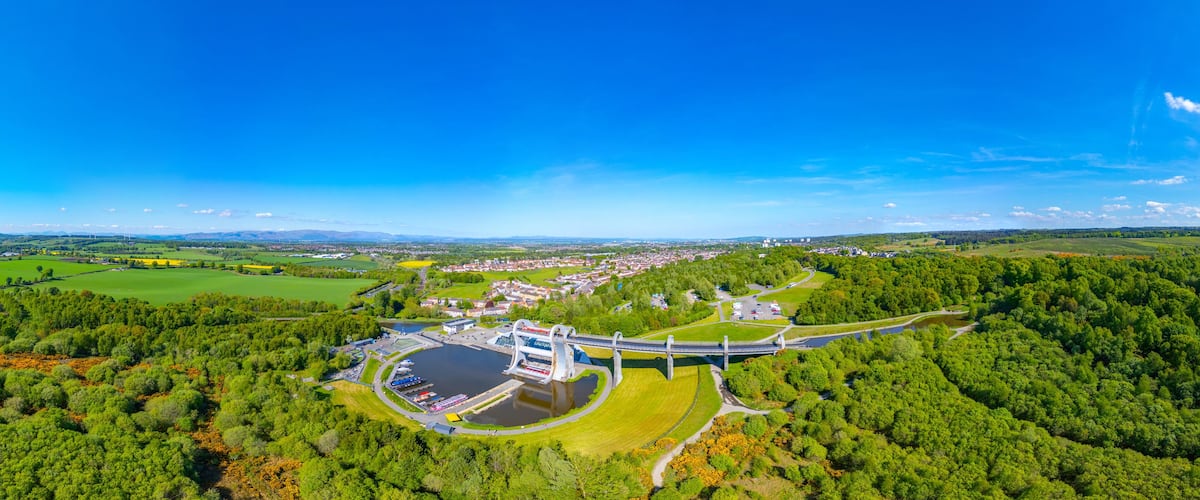 A panoramic view of a town with a distinctive bridge, lush green fields, and a clear blue sky.