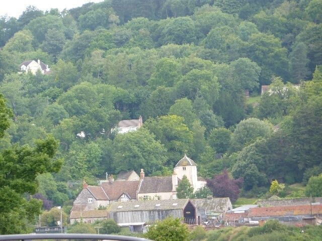 A village nestled in a valley with a prominent church and several houses surrounded by dense greenery.