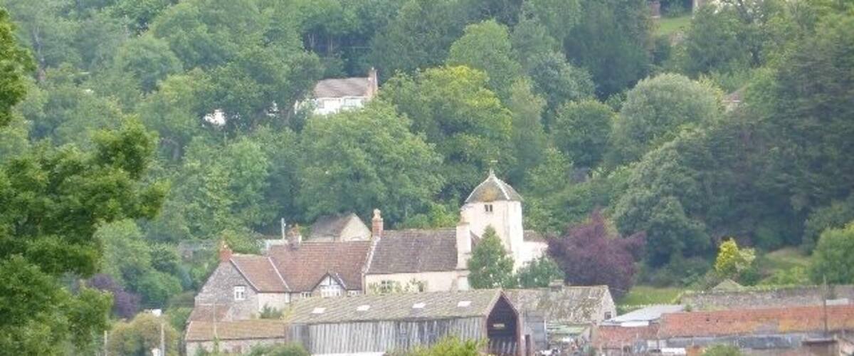 A village nestled in a valley with a prominent church and several houses surrounded by dense greenery.