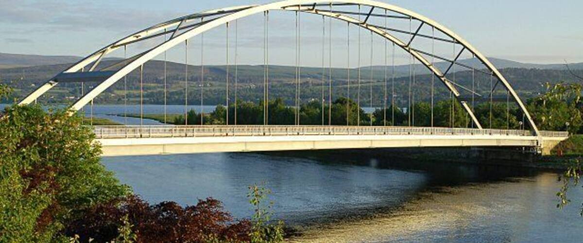 Een witte, gebogen brug over een rivier, met heuvels op de achtergrond.