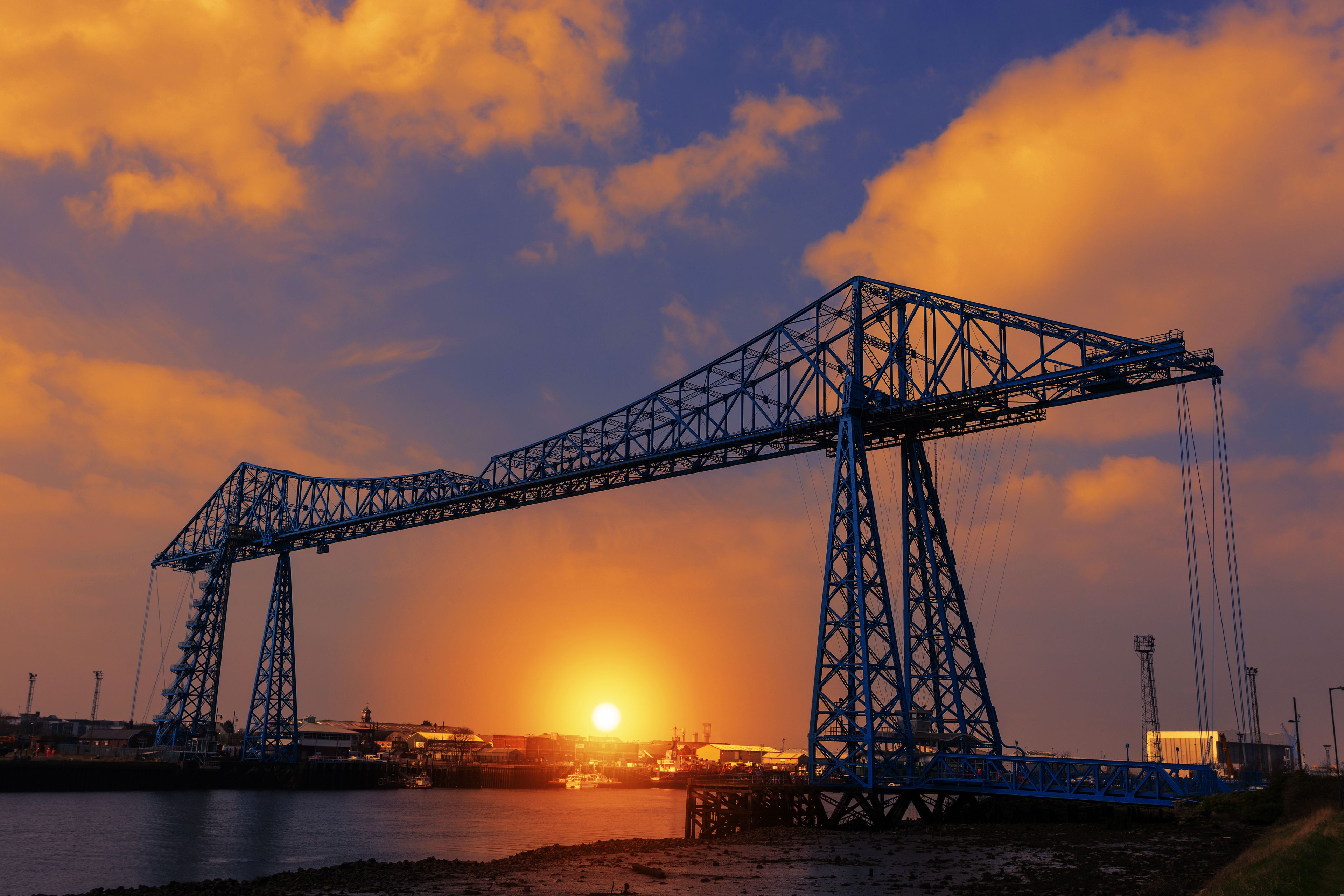 Uma grande ponte de aço atravessa um corpo de água, com o sol se pondo ao fundo.