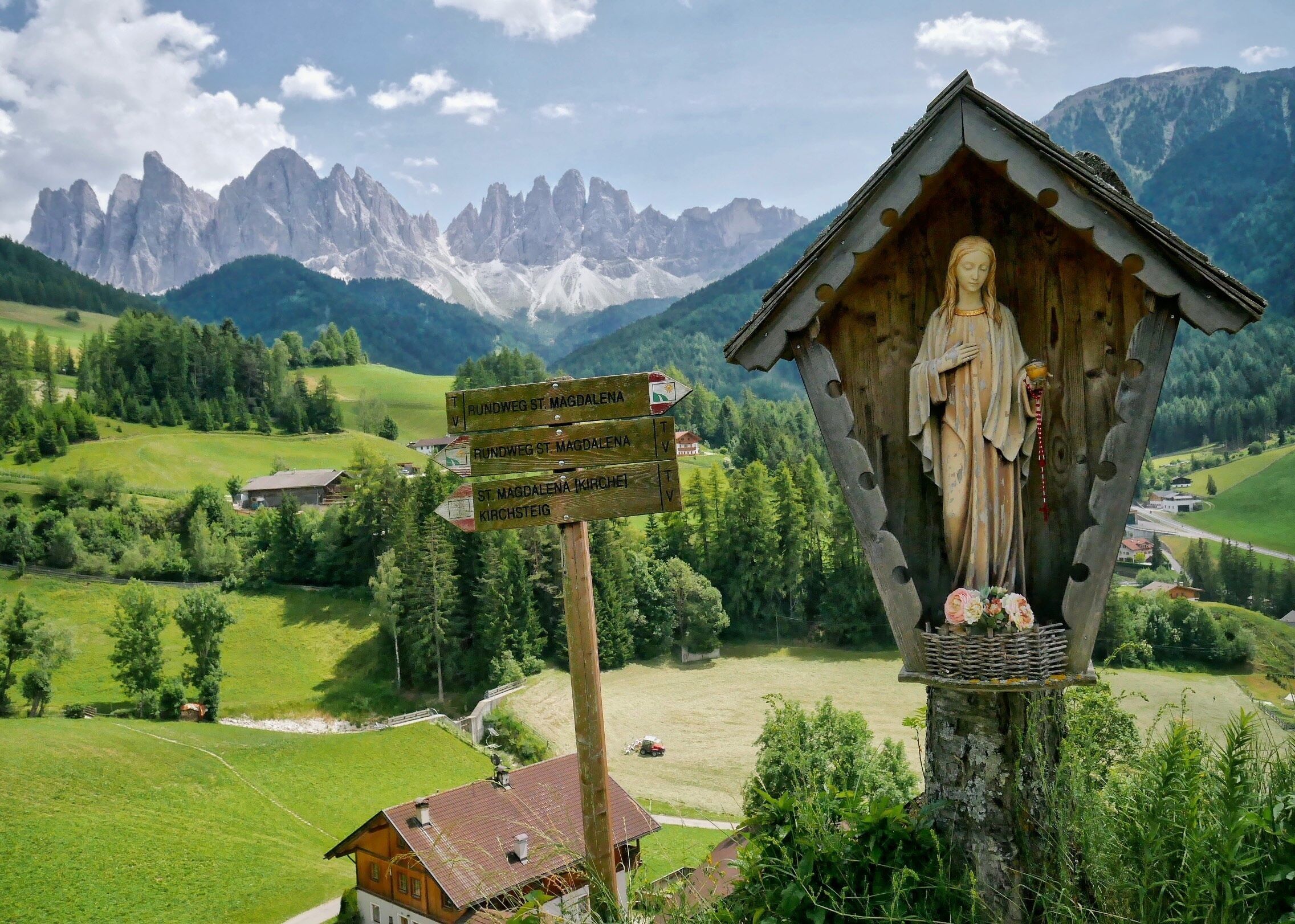 A wooden chapel with a statue of a woman, surrounded by a mountainous landscape and directional signs.