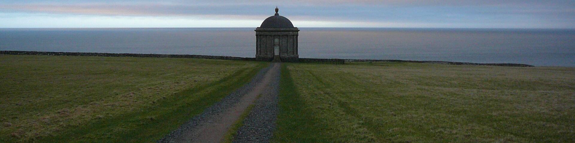 A solitary building with a dome on a grassy field, leading to a body of water.