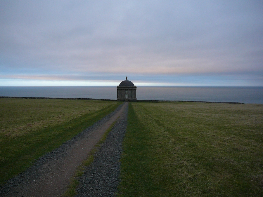 A solitary building with a dome on a grassy field, leading to a body of water.