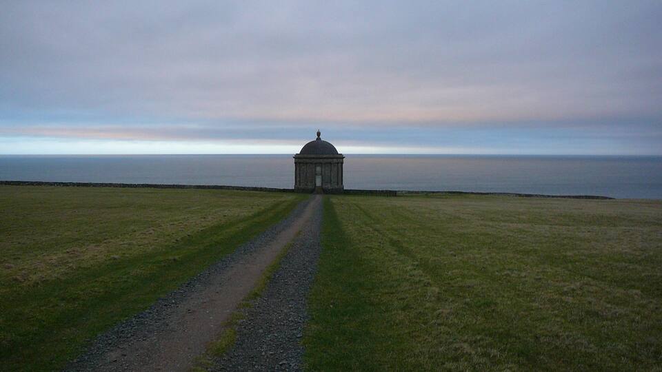A solitary building with a dome on a grassy field, leading to a body of water.