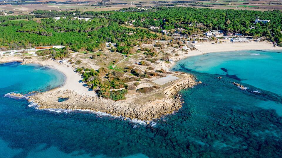 An aerial view of a coastal area with a sandy beach, clear turquoise waters, rocky shoreline, and a forested hill in the background.