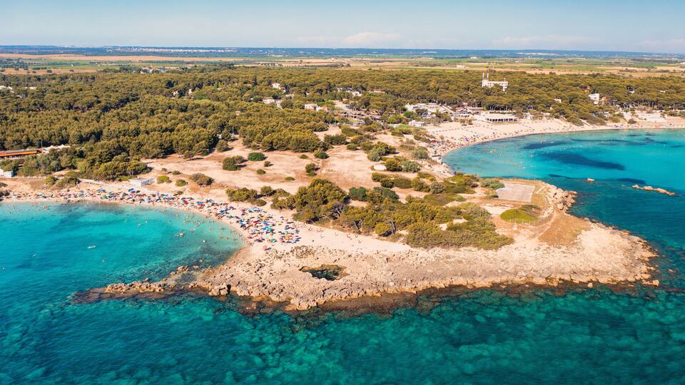 An aerial view of a coastal area with clear turquoise waters, a sandy beach, and a dense forested area.