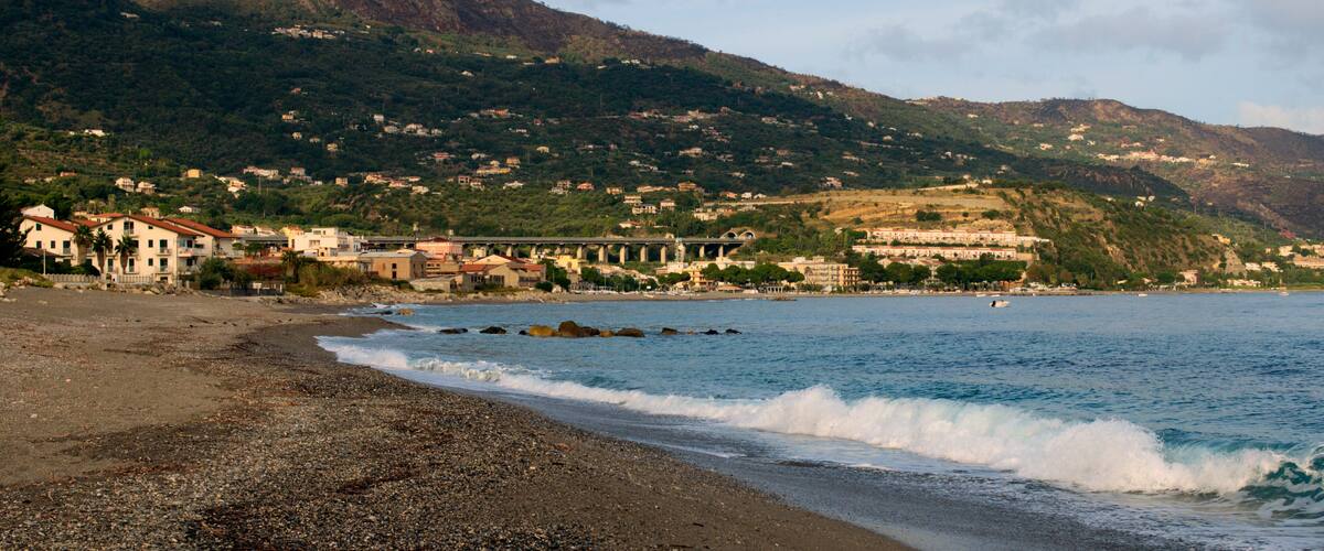 A pebble beach with waves, a coastal town, and a mountainous backdrop.