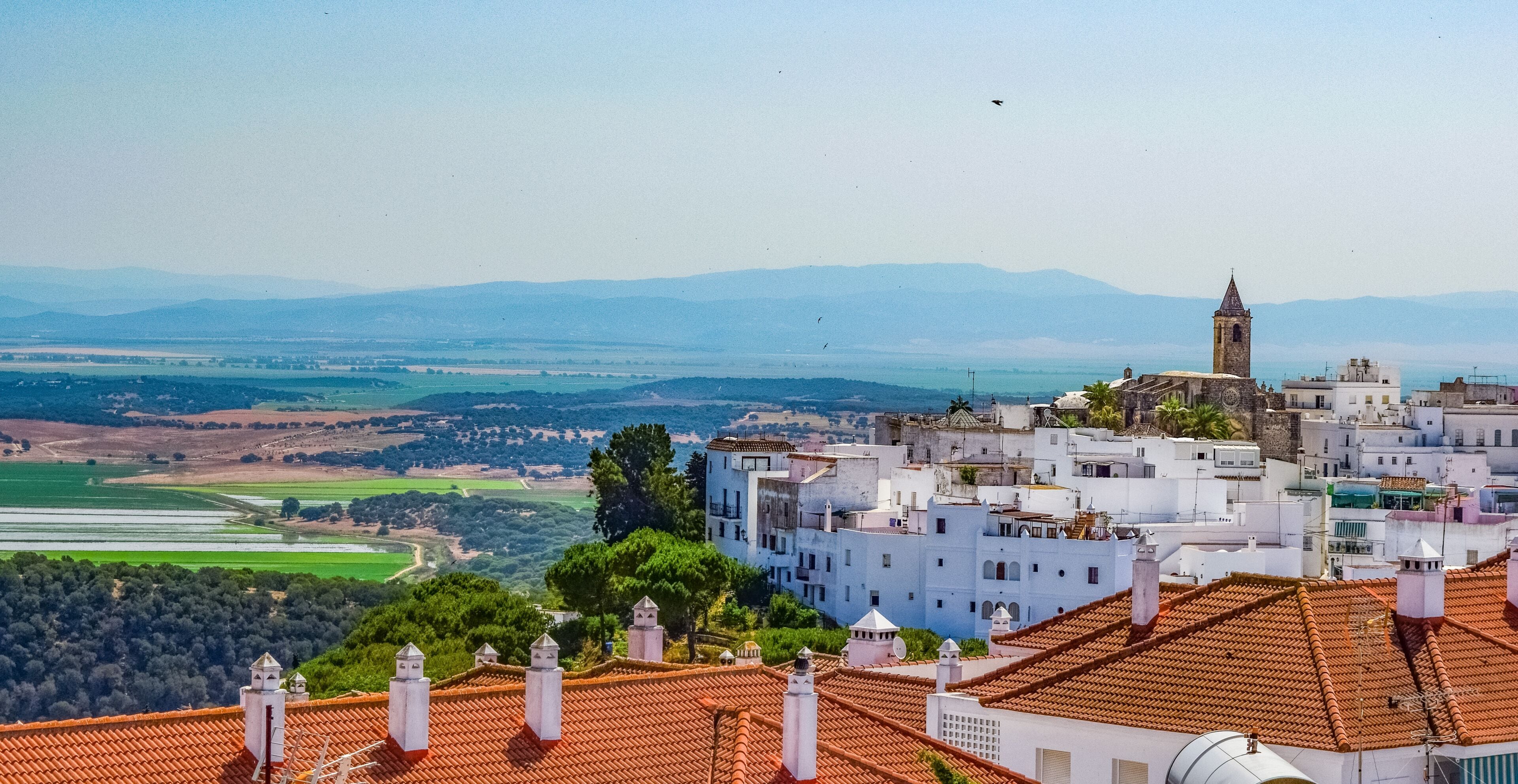A coastal town with white buildings, a prominent tower, terracotta rooftops, and a view of the sea and distant mountains.