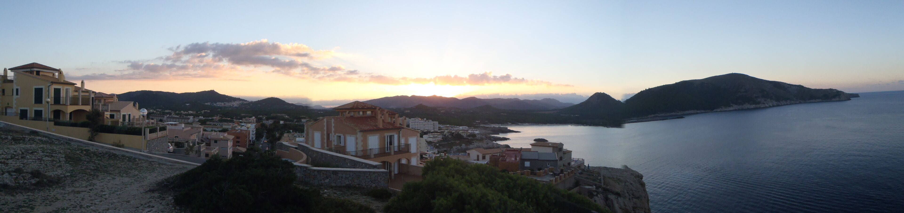 A coastal town with Mediterranean architecture, a calm sea, and a sunset over the hills.