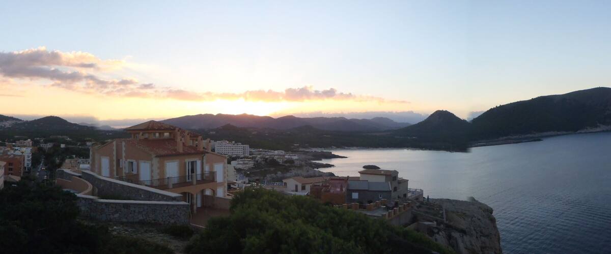 A coastal town with Mediterranean architecture, a calm sea, and a sunset over the hills.