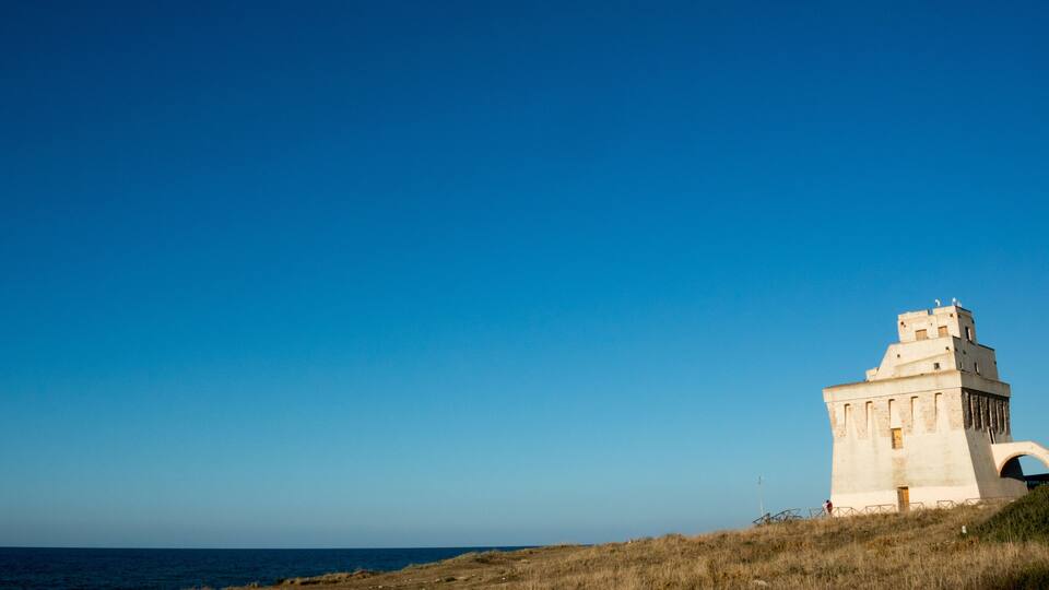 A coastal landscape with a prominent white tower-like structure on a grassy hill, overlooking the sea.