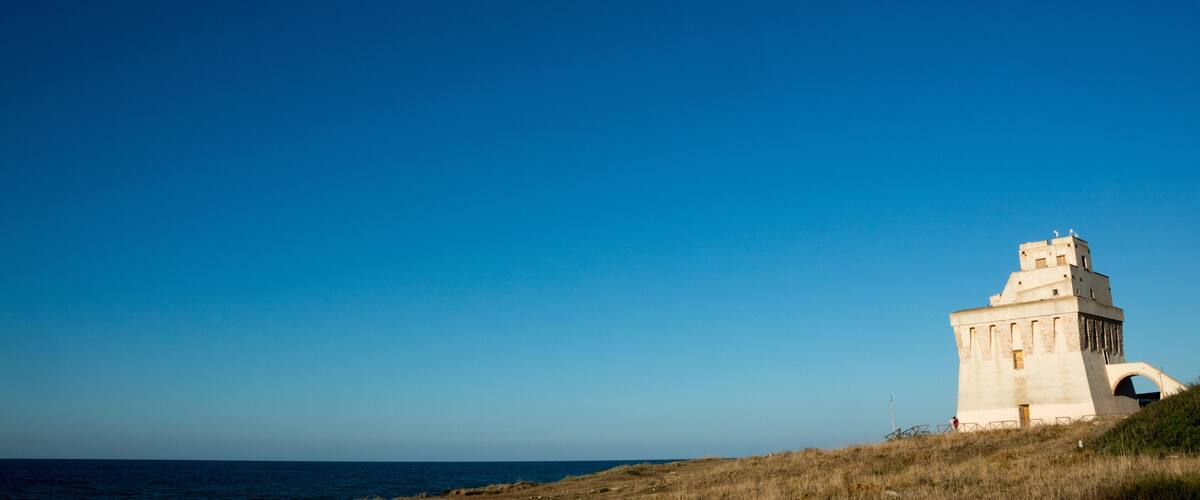 A coastal landscape with a prominent white tower-like structure on a grassy hill, overlooking the sea.