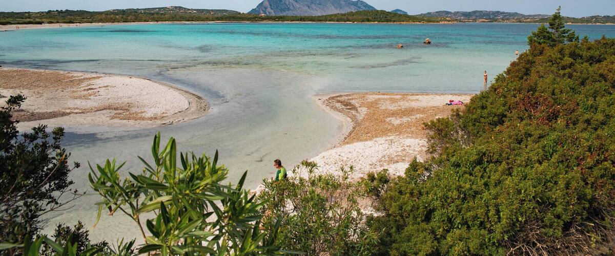A coastal landscape with clear turquoise waters, sandy beaches, and a prominent mountain in the background.