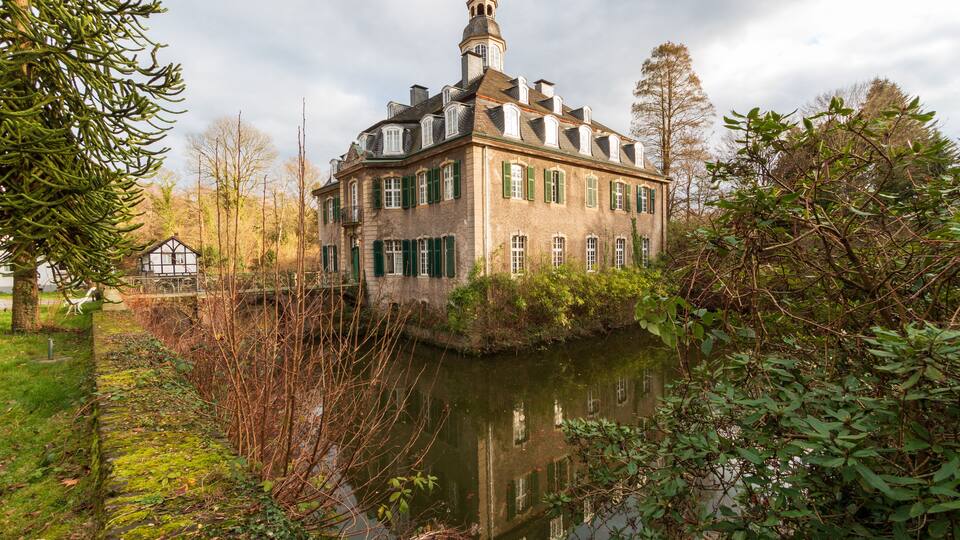 Un edificio histórico con una torre, rodeado por un río y abundante vegetación.