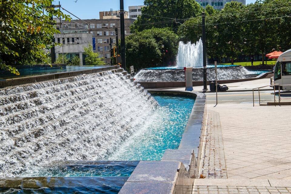 A water feature with cascading water, a paved walkway, and buildings in the background.
