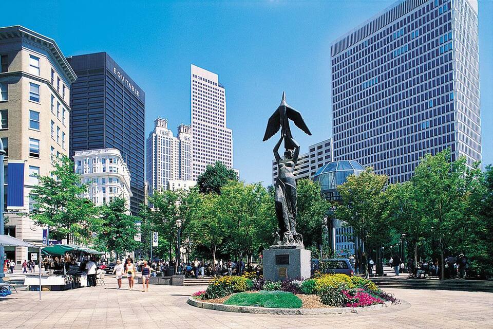 Una plaza urbana con una estatua de una persona sosteniendo un paraguas, rodeada de árboles y flores.