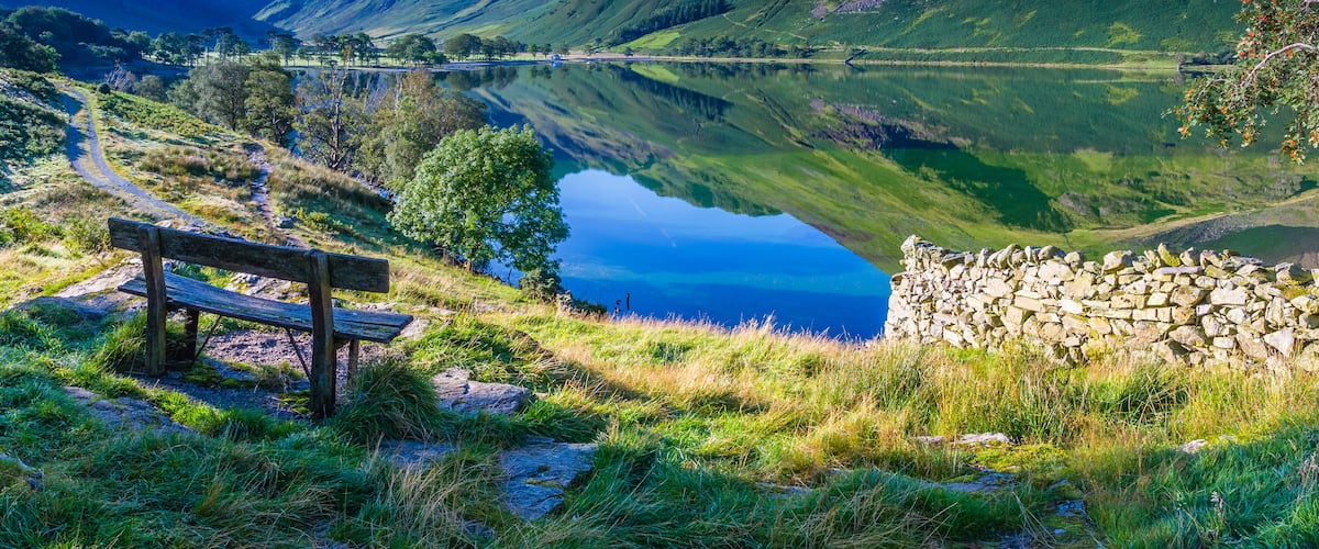 A tranquil lake surrounded by mountains, a wooden bench, and a stone wall.