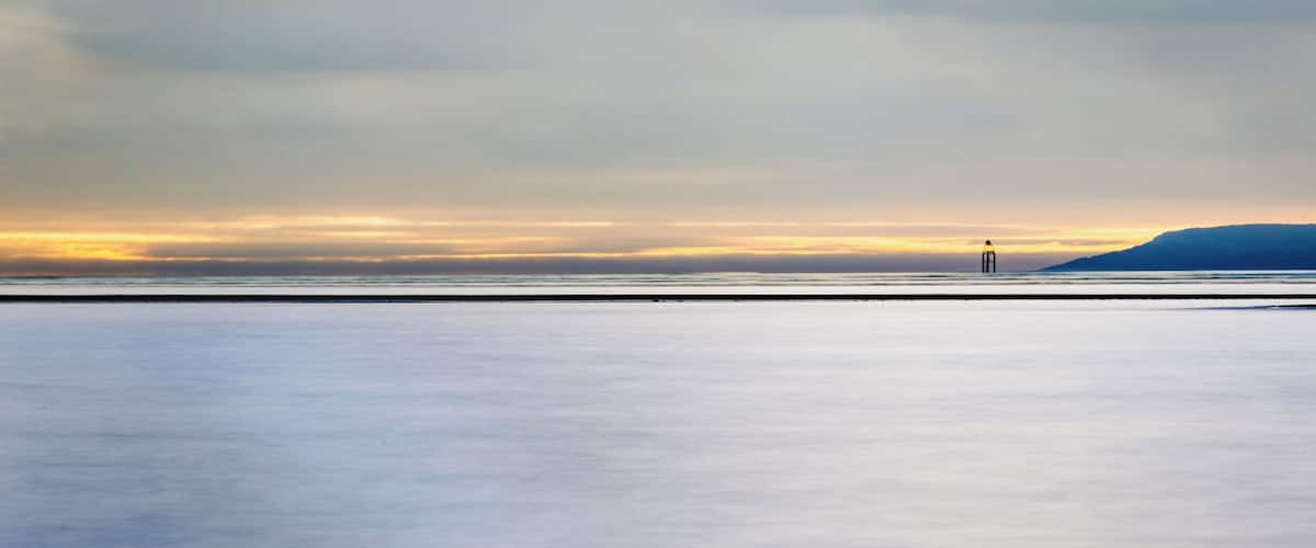 A calm sea at sunset with a distant lighthouse and a hilly landscape.
