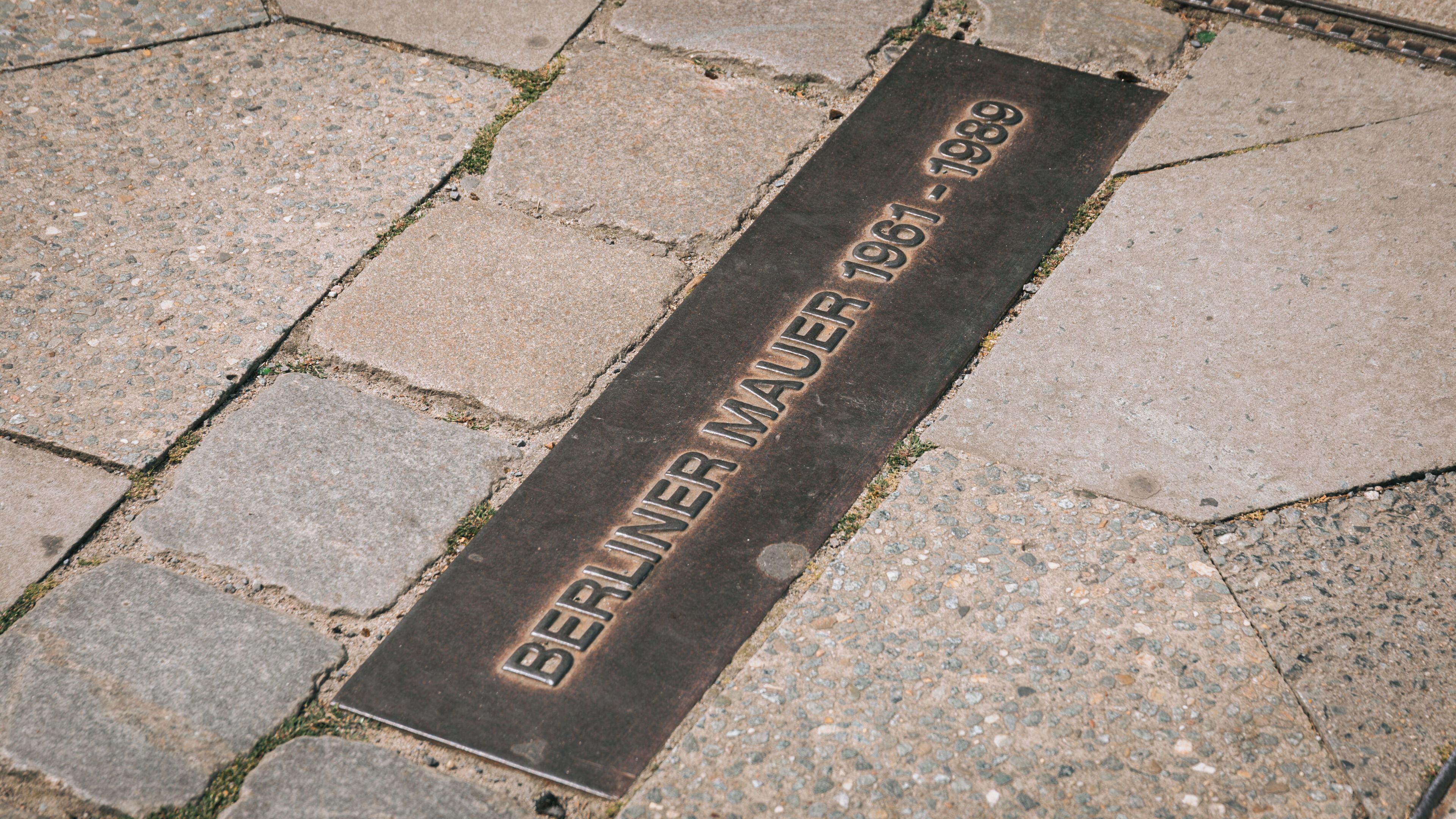 Une plaque métallique portant l’inscription « BERLIN MAUER 1961-1989 » est fixée sur une surface pavée.