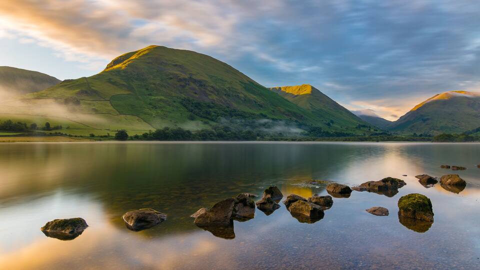 A calm lake with rocks in the foreground, surrounded by green hills and mountains under a cloudy sky.