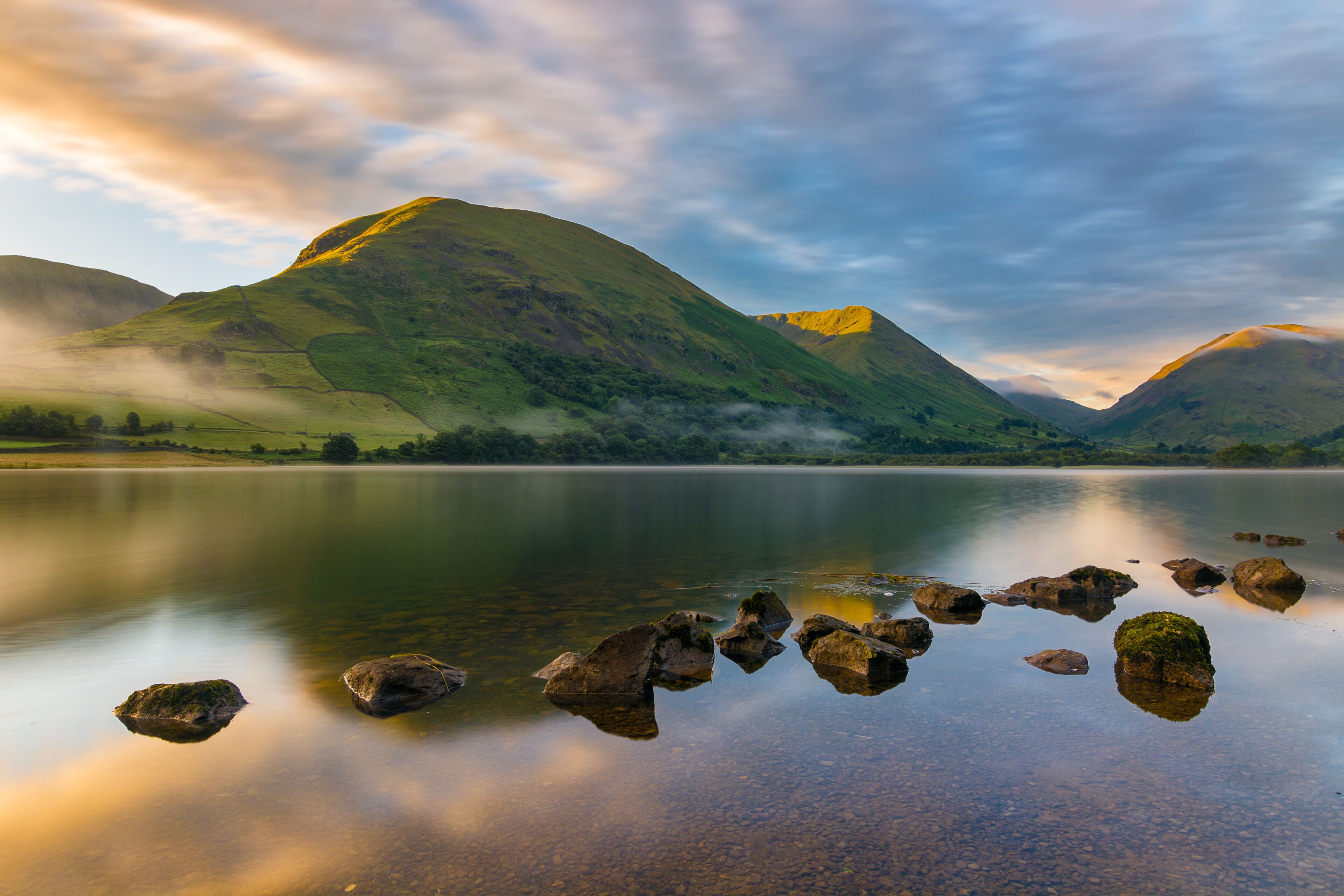 A calm lake with rocks in the foreground, surrounded by green hills and mountains under a cloudy sky.
