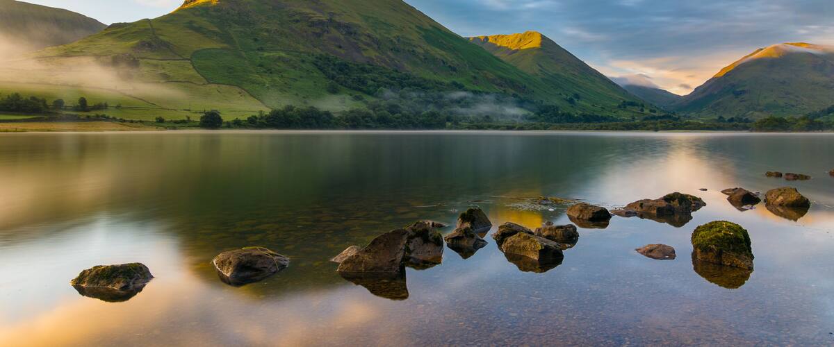 A calm lake with rocks in the foreground, surrounded by green hills and mountains under a cloudy sky.