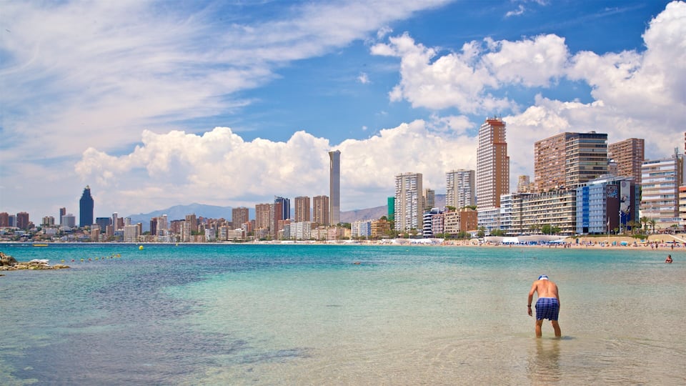 A beach with clear turquoise water, a city skyline with tall buildings, and a person standing in the water.
