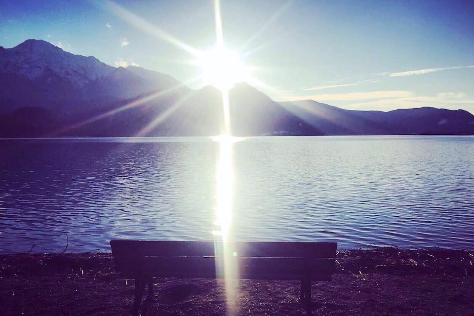 A bench facing a calm lake with mountains in the background and the sun shining brightly.