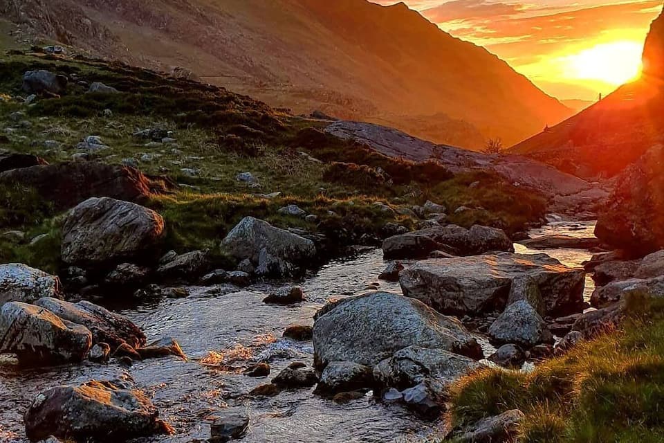 A mountain stream with rocks, surrounded by grassy terrain and mountains during sunset.