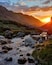 A mountain stream with rocks, surrounded by grassy terrain and mountains during sunset.