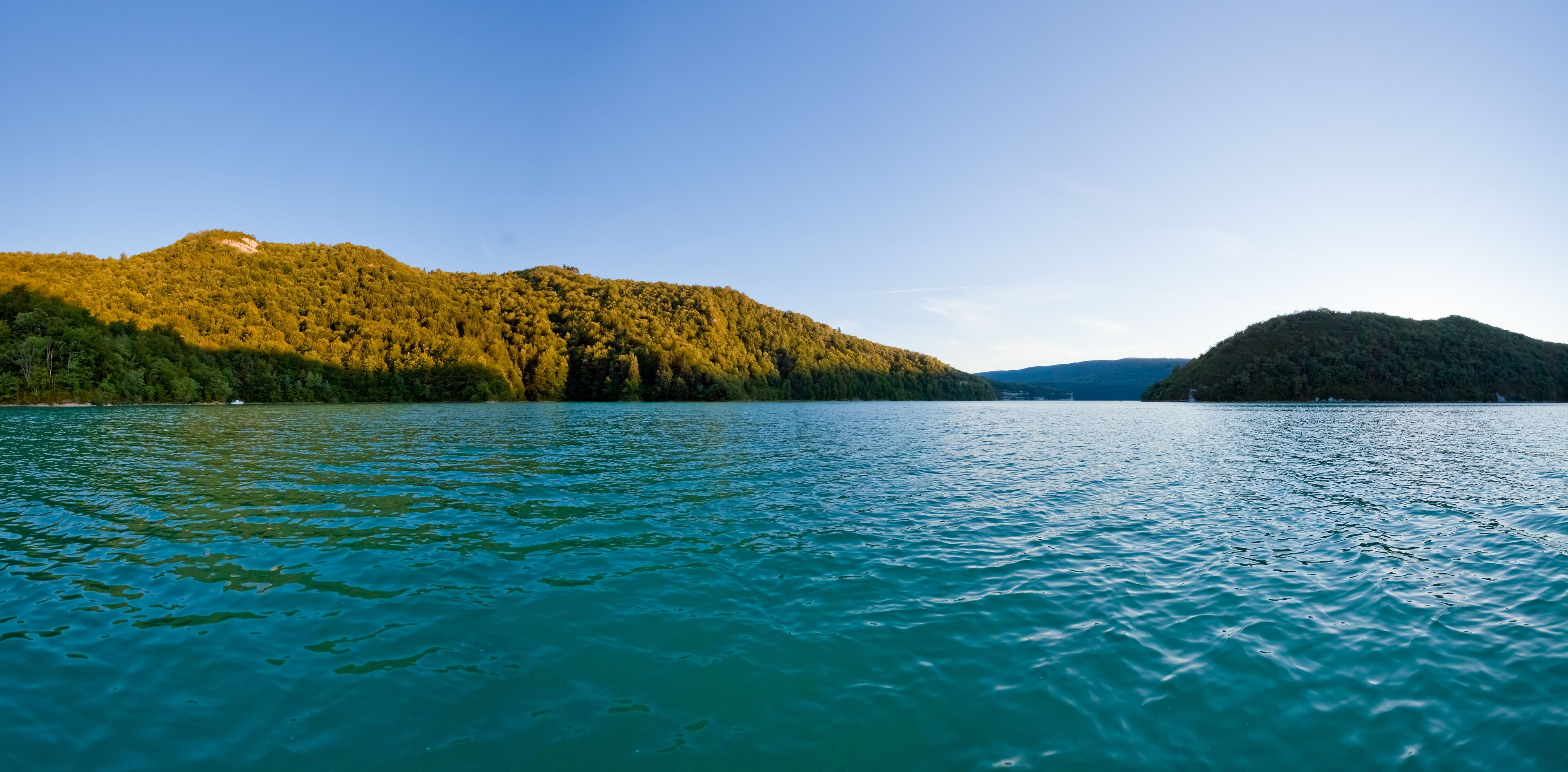 A calm lake with clear turquoise water, surrounded by forested hills under a clear sky.