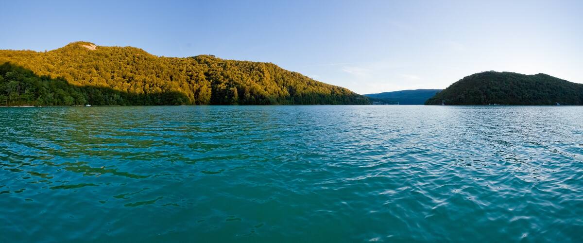 A calm lake with clear turquoise water, surrounded by forested hills under a clear sky.