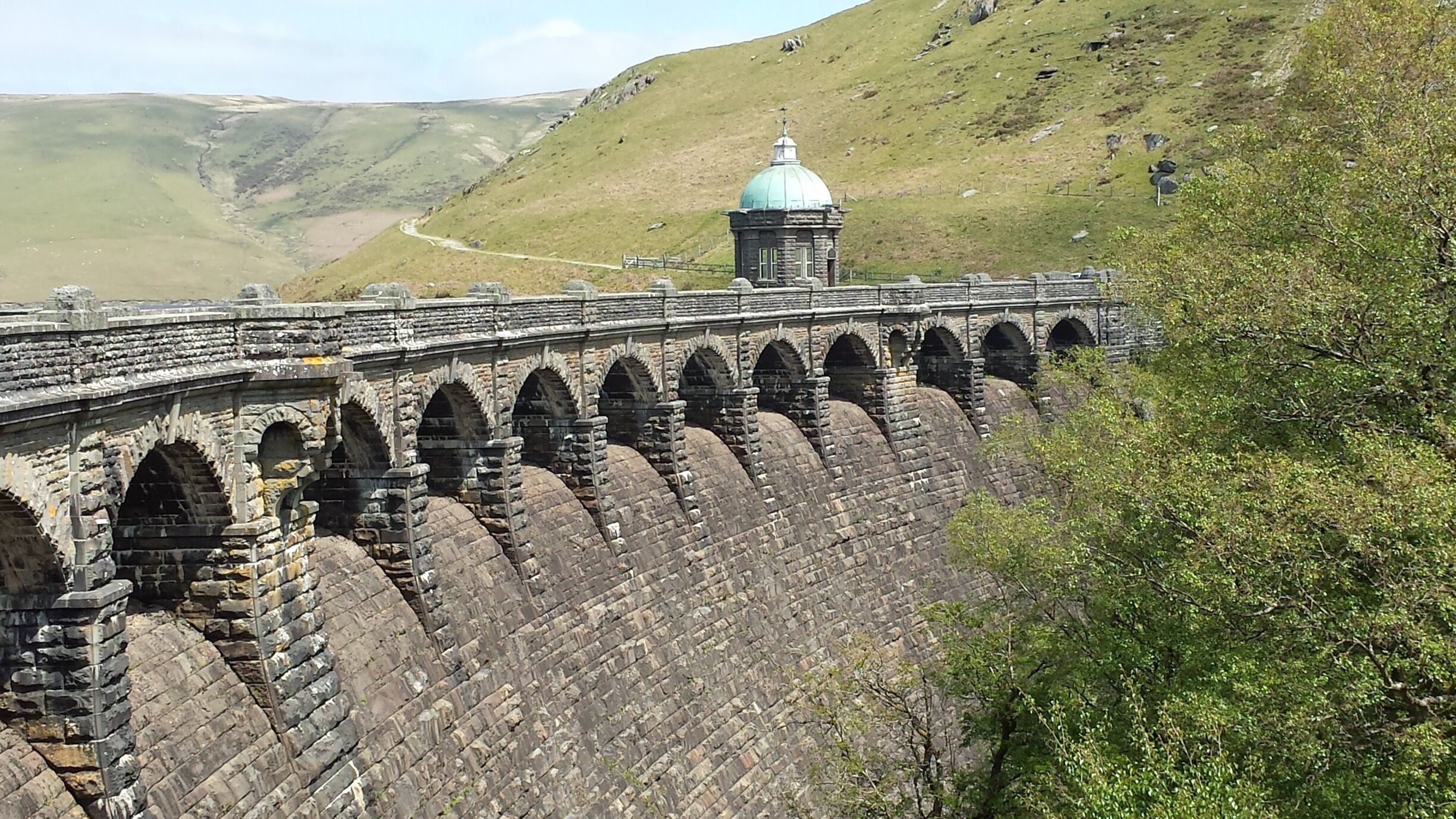 A stone viaduct with multiple arches spanning a valley, a small building with a dome, and a winding road in the background.