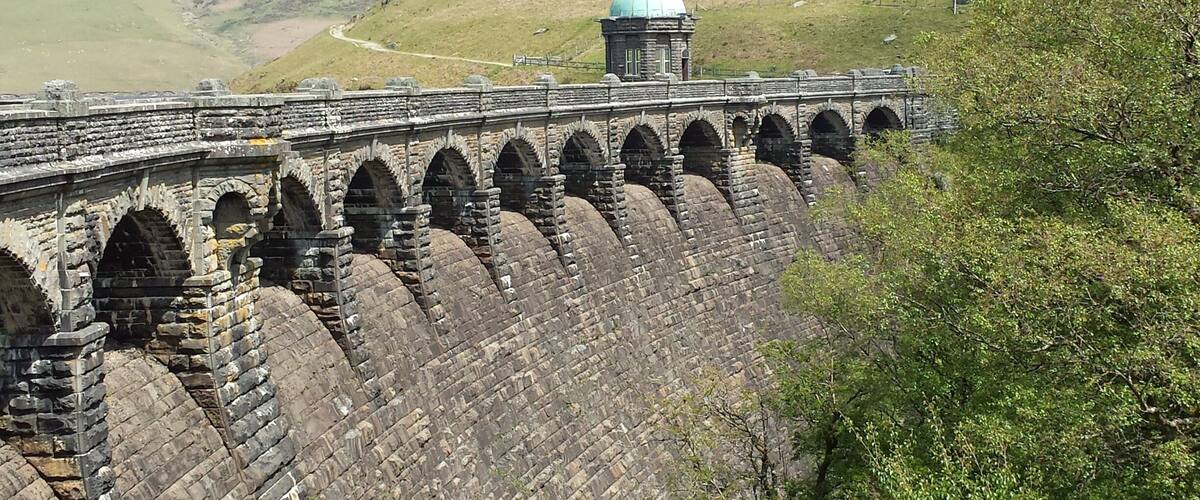 A stone viaduct with multiple arches spanning a valley, a small building with a dome, and a winding road in the background.