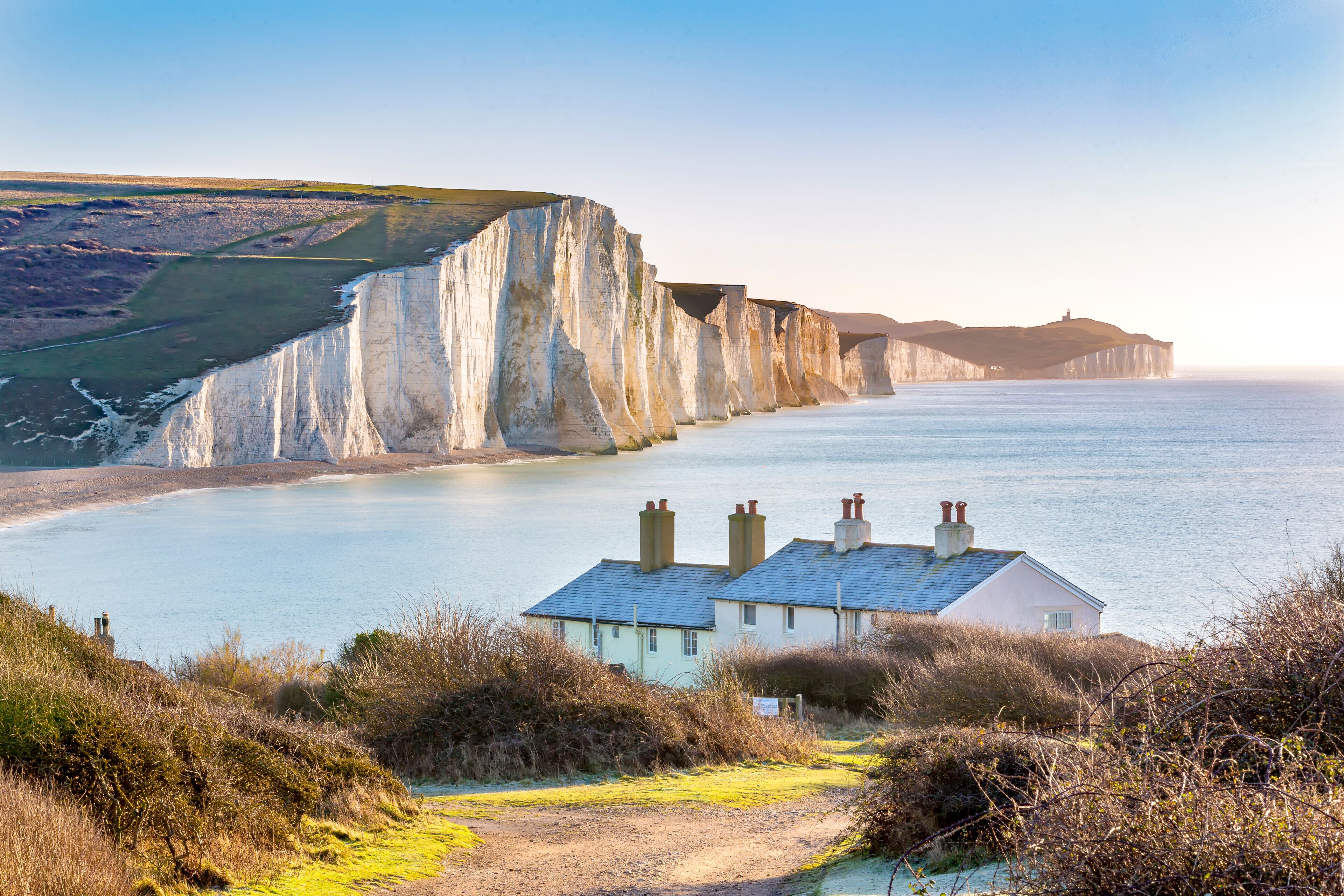A coastal landscape with white cliffs, a calm sea, and a house with chimneys.