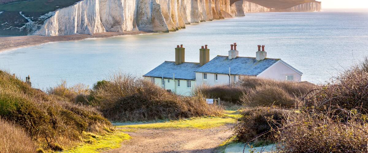 A coastal landscape with white cliffs, a calm sea, and a house with chimneys.