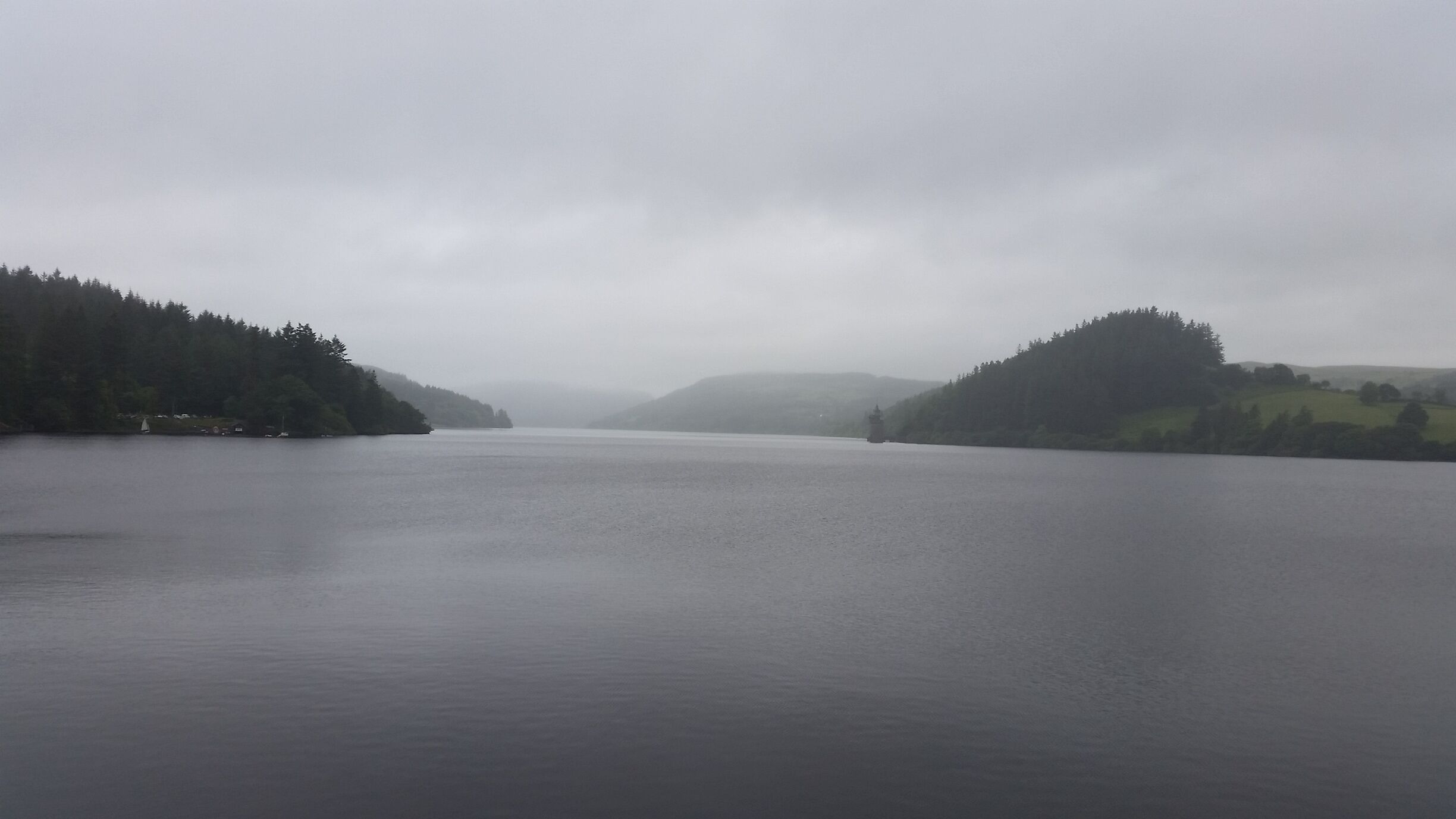 A calm lake surrounded by forested hills under a cloudy sky.
