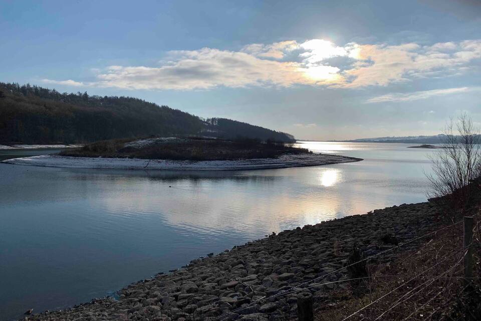 A calm river with a rocky shoreline, surrounded by forested hills and a clear sky.