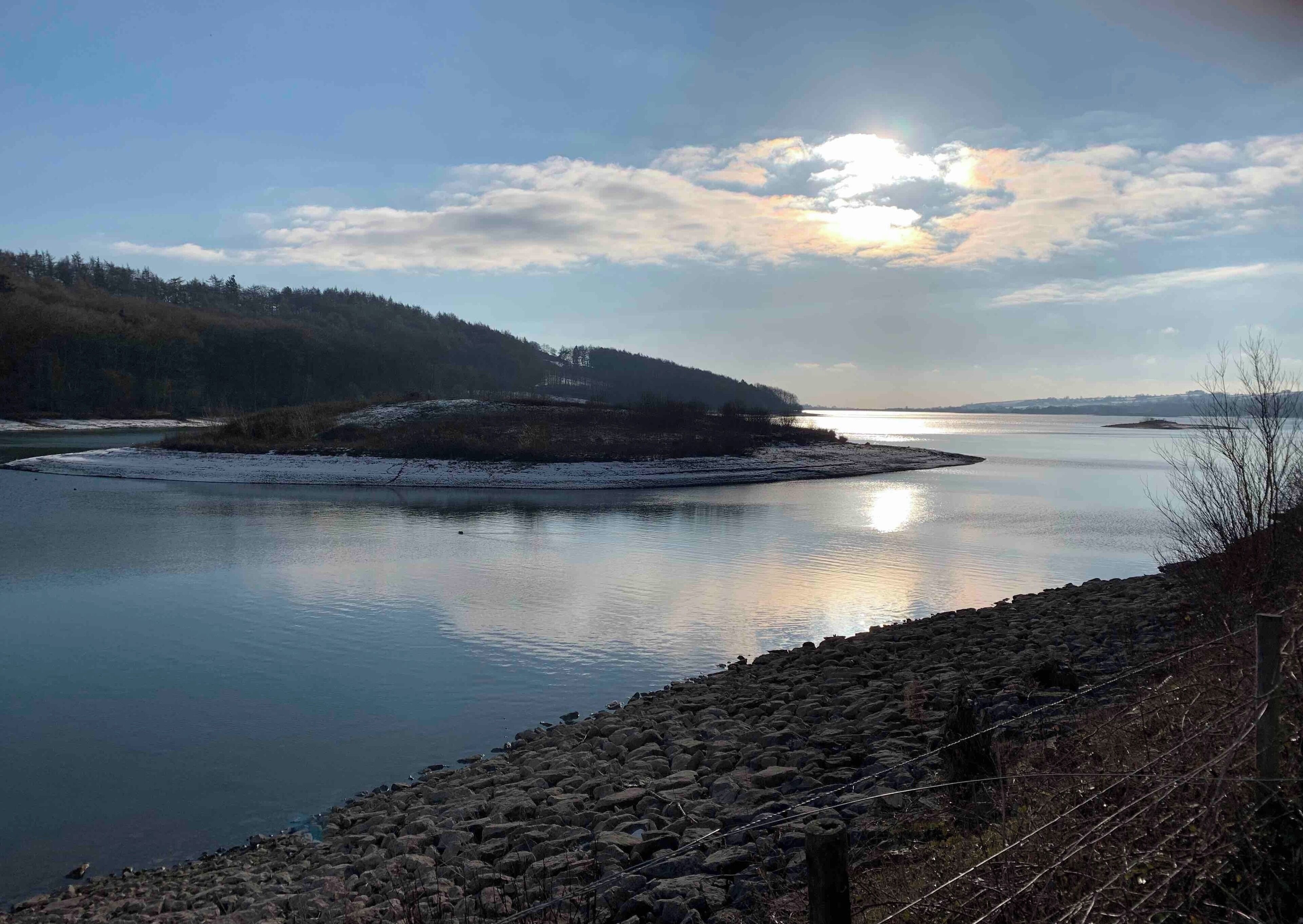 A calm river with a rocky shoreline, surrounded by forested hills and a clear sky.