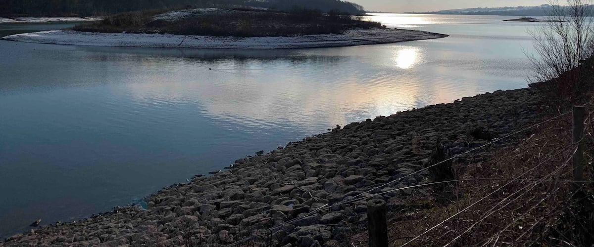 A calm river with a rocky shoreline, surrounded by forested hills and a clear sky.