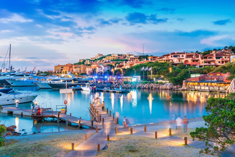 A marina with yachts, a wooden jetty, and buildings on a hillside.