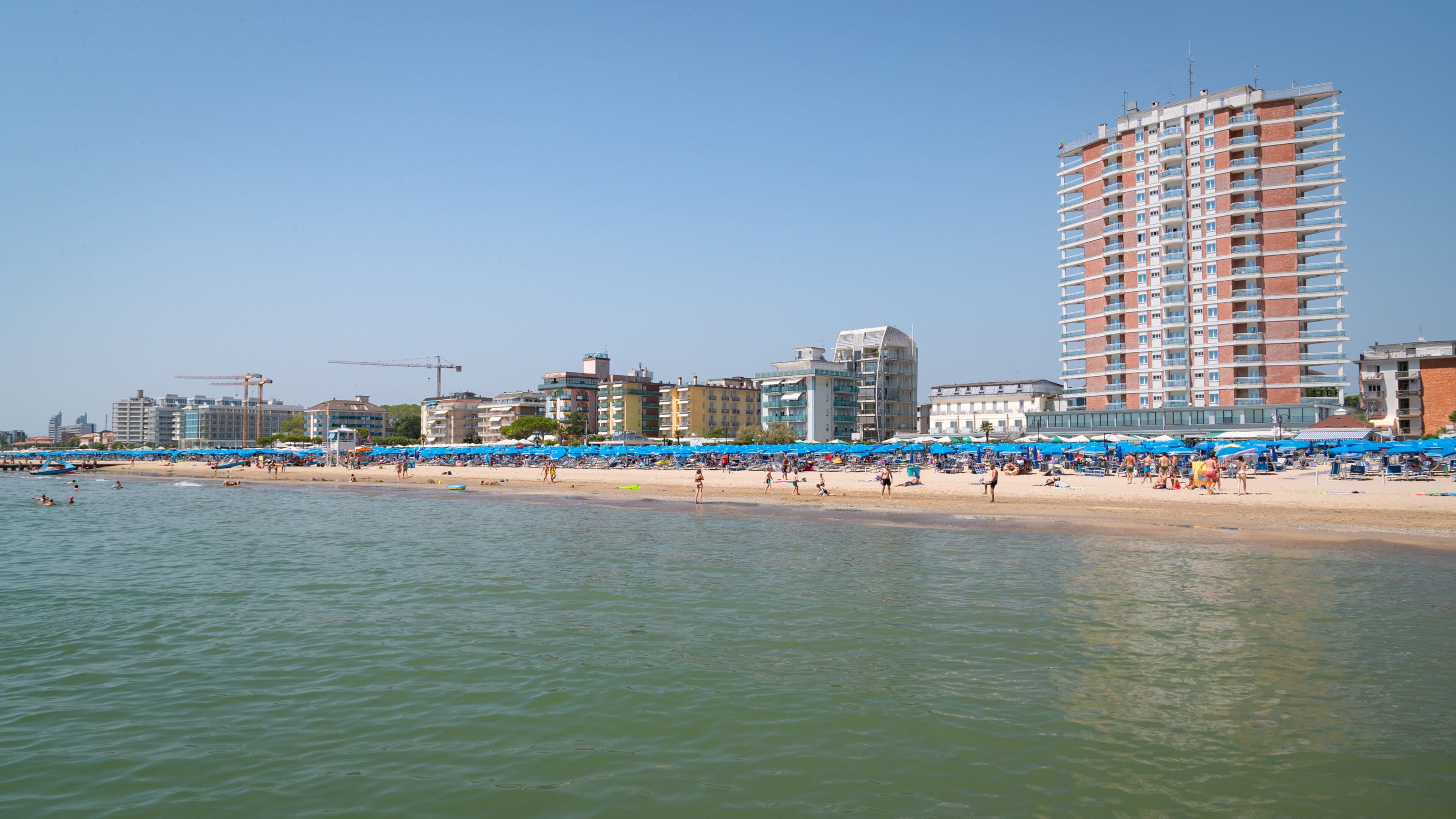 A beach with blue umbrellas and people. There are buildings and a tall hotel in the background.