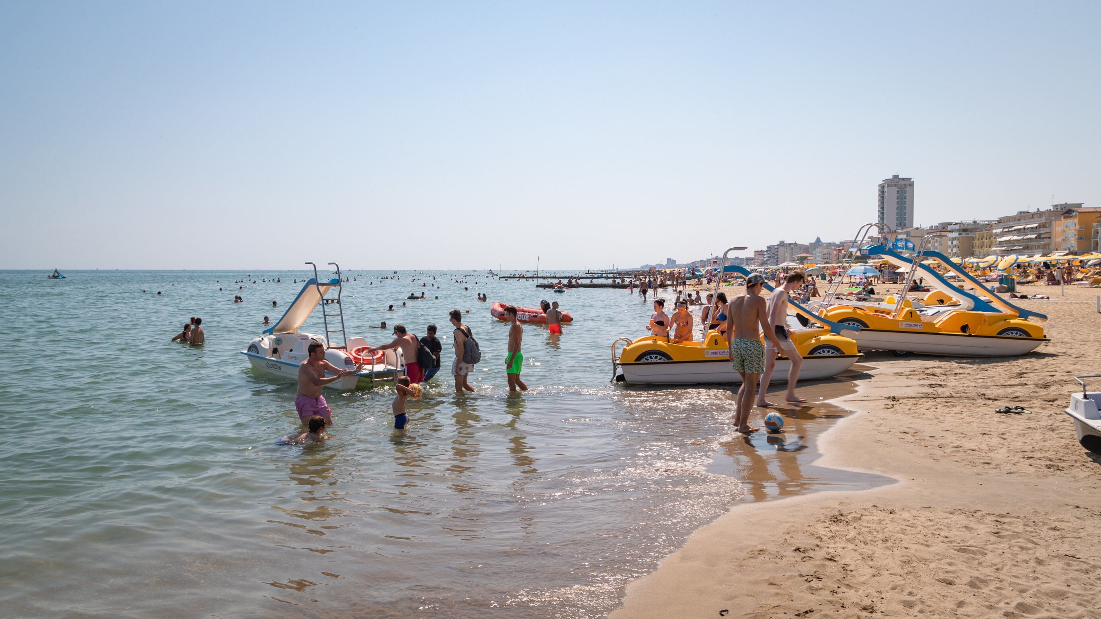 A crowded beach with people swimming and standing in the water, and several yellow and white boats parked on the sand.