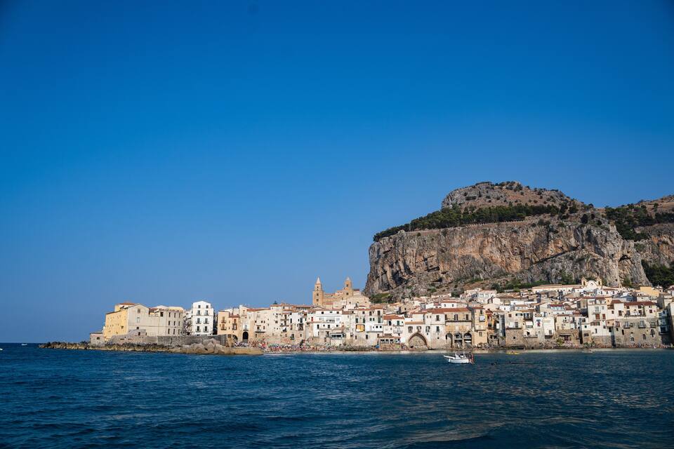 A coastal town with buildings along the shore, a prominent cliff, and a small boat in the water.