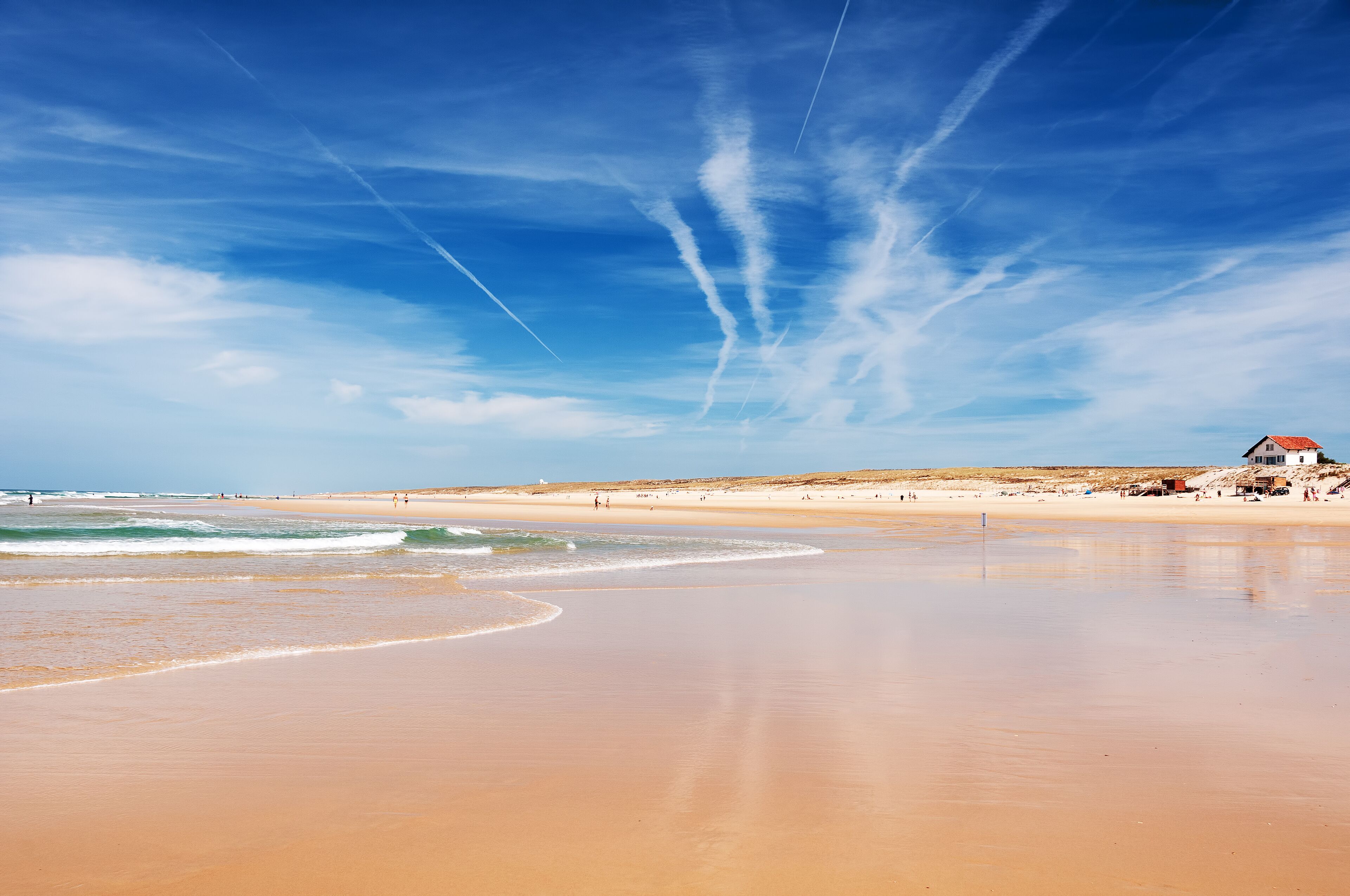 A wide sandy beach with gentle waves, a clear blue sky, and contrails.